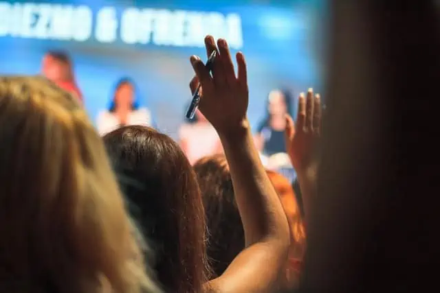 A shot from the crowd up to the stage. Multiple people are raising their hand.